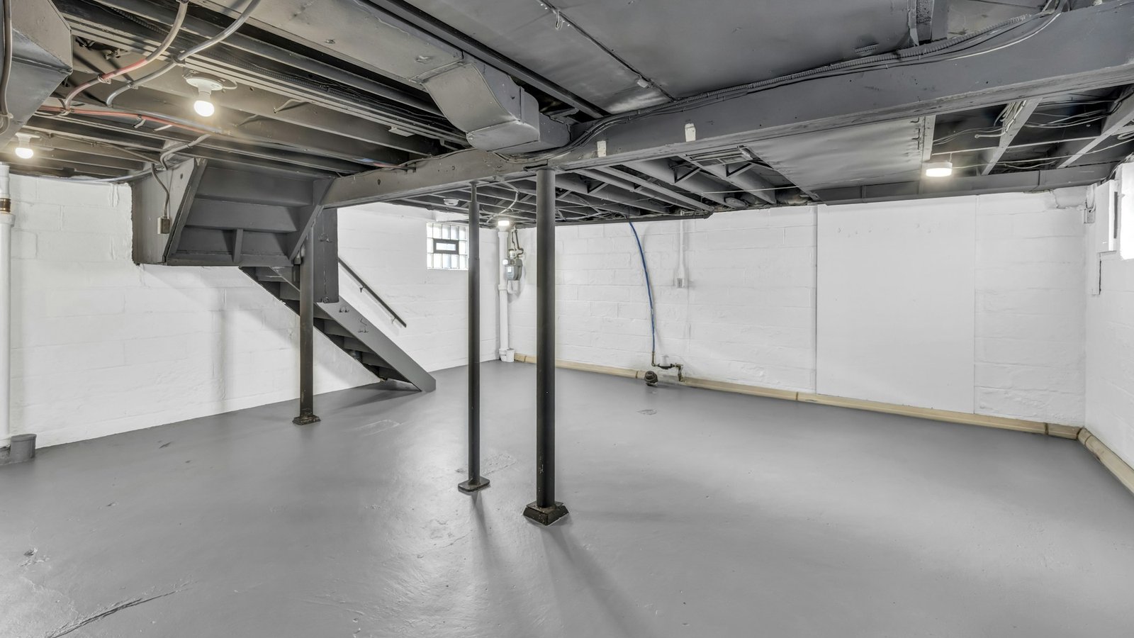 Unfinished basement with exposed joists, steel support poles and a staircase, before framing
