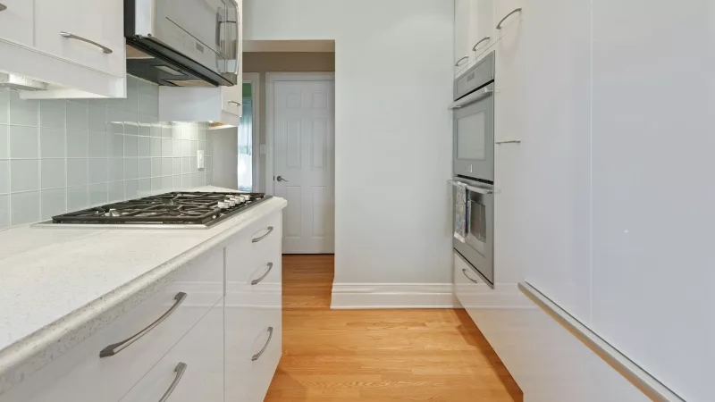 Narrow galley kitchen with white cabinets, a gas cooktop, wall ovens and a hardwood floor leading to a doorway