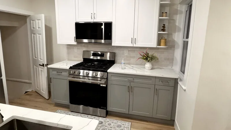 Remodeled rowhome kitchen with white upper and gray lower shaker cabinets, quartz counters and a stainless gas range