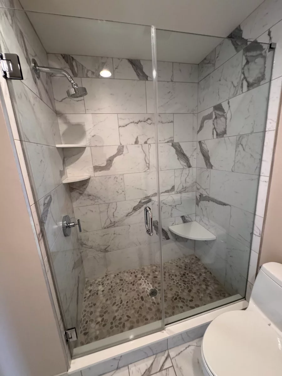 Walk-in shower with marble-look porcelain walls, corner shelves, pebble tile floor and a frameless glass door in Feasterville, PA