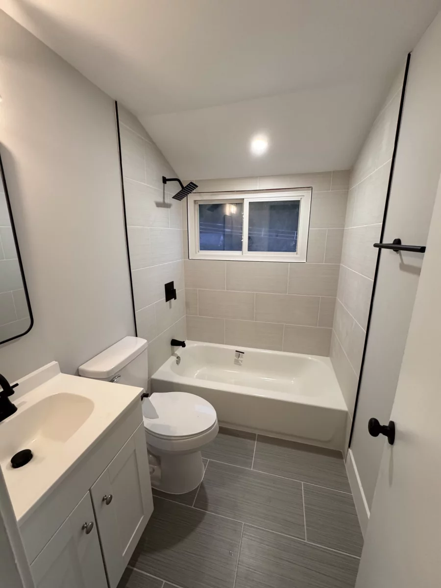 Bathroom with matte black fixtures, a white vanity, tub with tile surround and gray plank tile floor