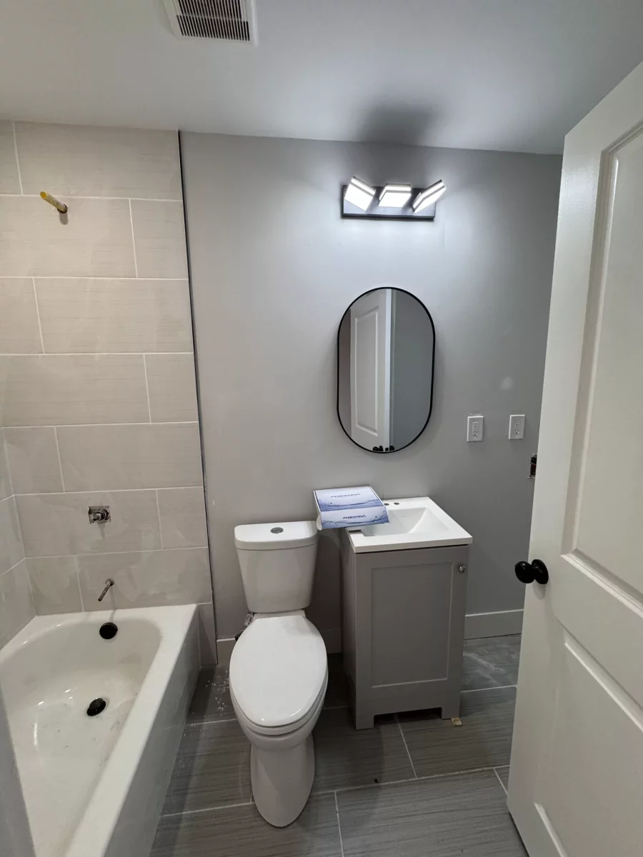 Bathroom with gray tile tub surround, gray plank floor, white vanity and oval mirror