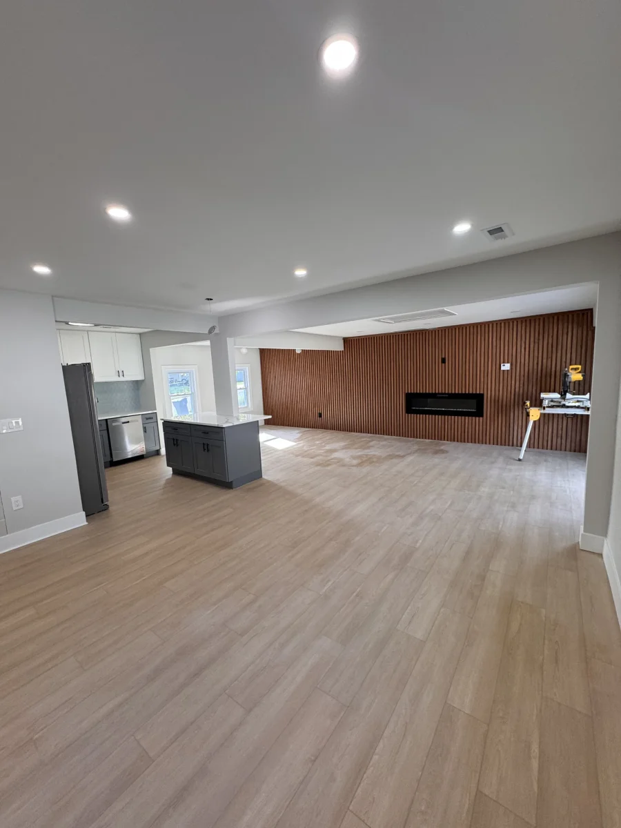 Open living area with light wood-look flooring, recessed lights, a kitchen island and a wood-slat wall with a linear fireplace in Levittown