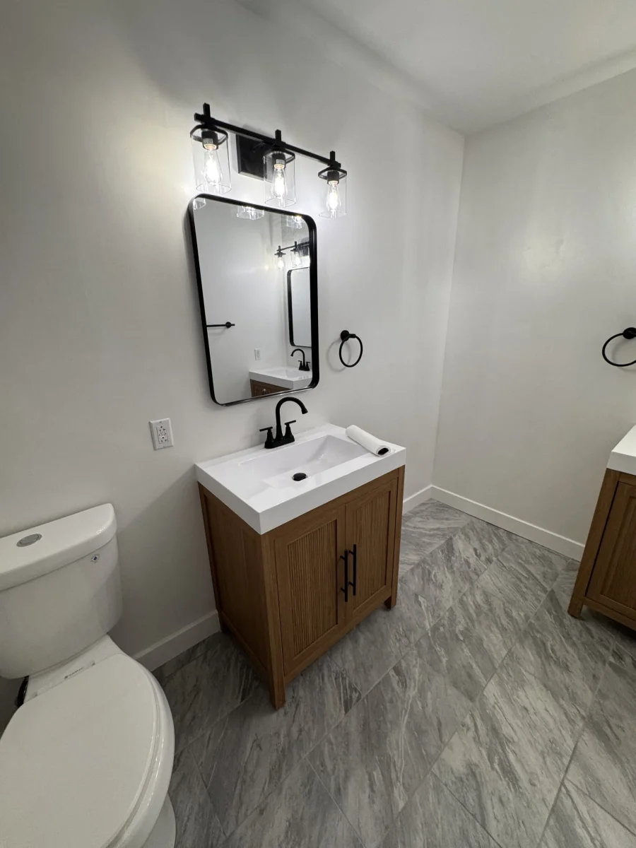 Wood vanity with a white top, black mirror and black vanity light over a gray tile floor