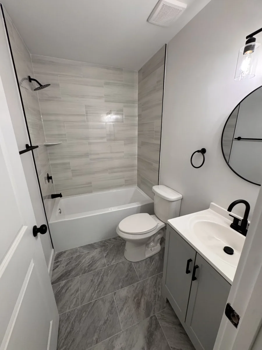 Bathroom with a tiled tub surround, gray vanity and round black mirror in Newtown